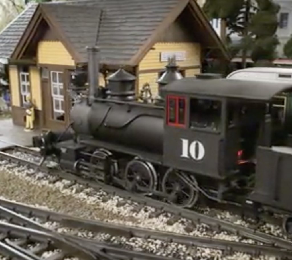 Rear left oblique view of a garden scale steam locomotive near a passenger depot.