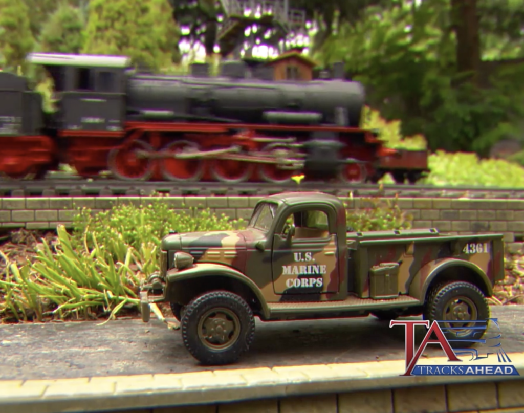 Garden scale, German-style steam locomotive passes behind a stationary U.S. Marines truck.