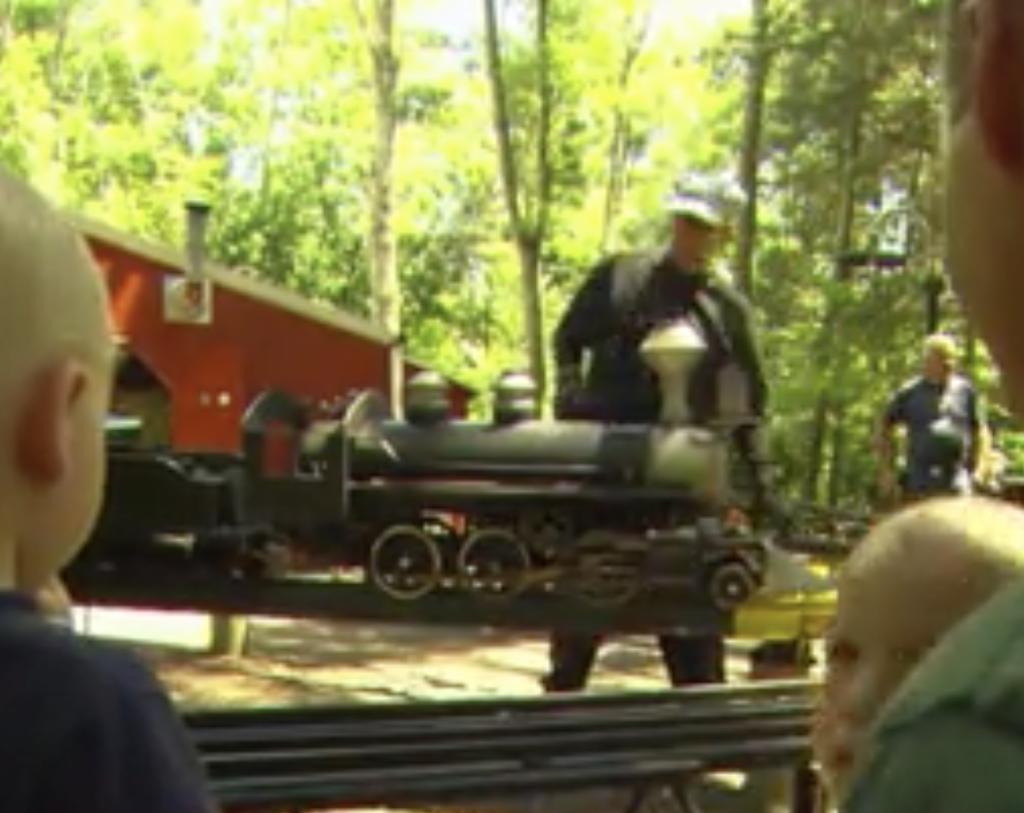 Children look on as a man works on a ride-on steam locomotive on an elevated platform.