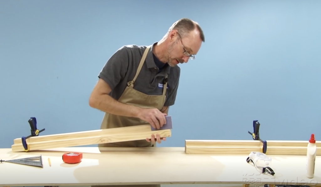 Man holding a table leg and sanding a piece of it in a workshop.