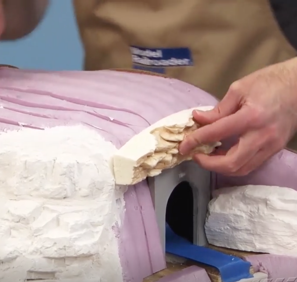 Hand placing foam "rocks" above a raw and unfinished tunnel portal on a model train layout.