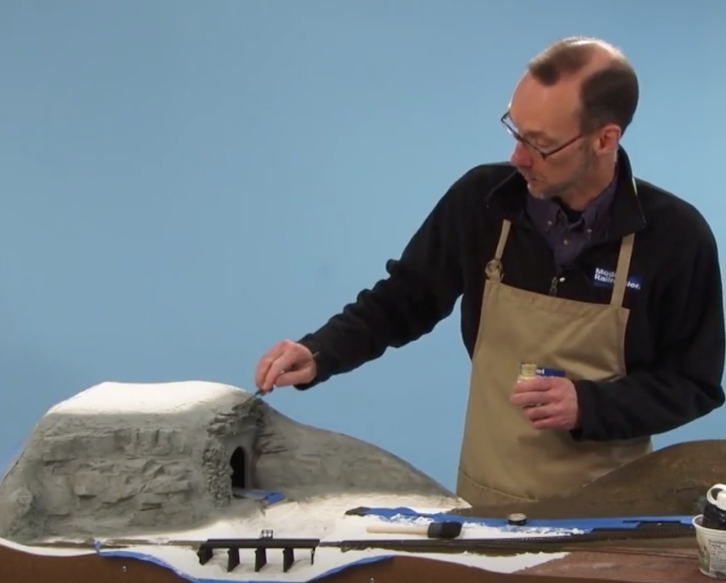 Man holding tool decorates "rocks" on a model train layout.