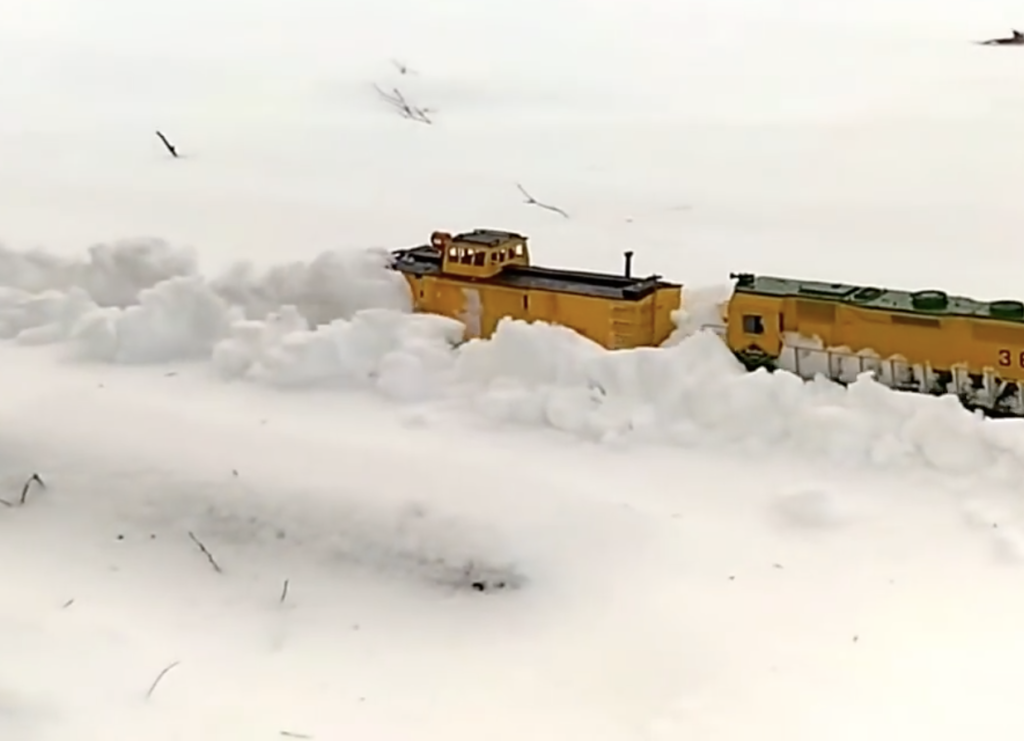 Yellow garden-scale railroad snowplow cleaves through snow on an outdoor layout.