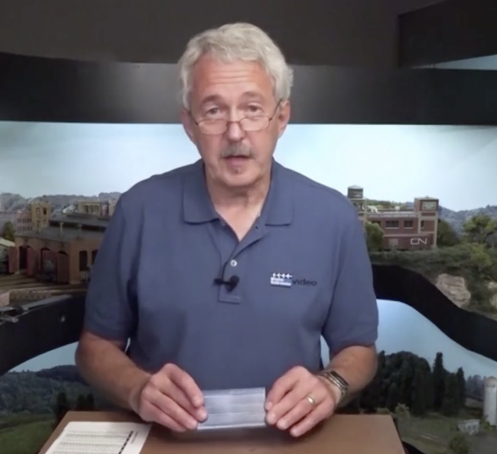 Man standing behind small table surrounded by model train layout holding small plastic box.