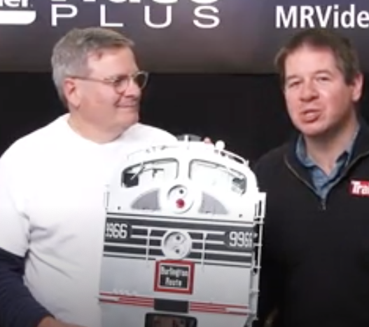 Two men standing with a sign shaped like a locomotive.