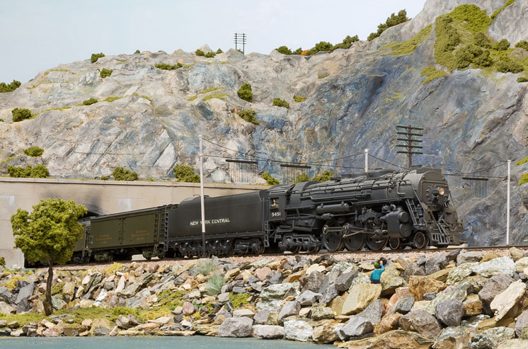 A model steam locomotive leads an express train through a mountainous scene.