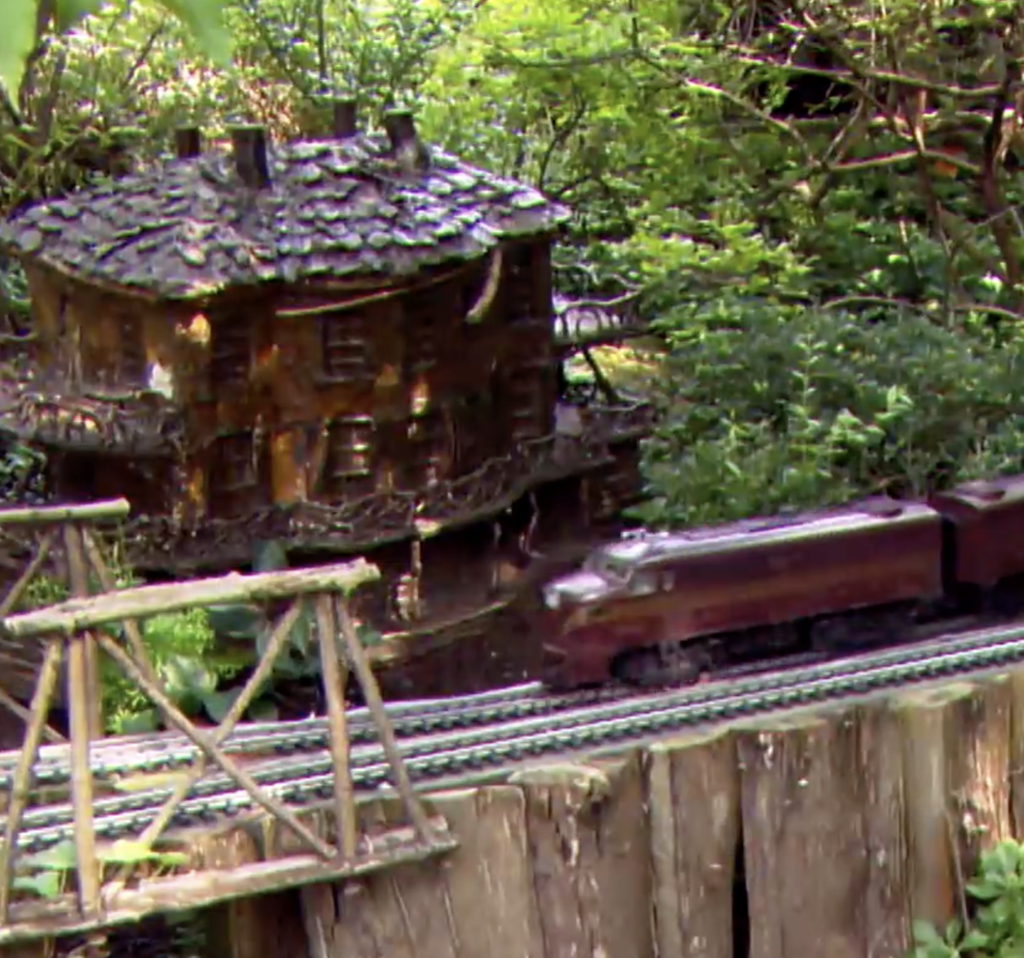 A garden-scale streamlined diesel locomotive leads a train beside a scale storybook architecture structure.