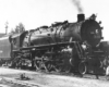 A black and white photo of locomotive 4-6-4 stopped at the rail yard as a person walks across the tracks