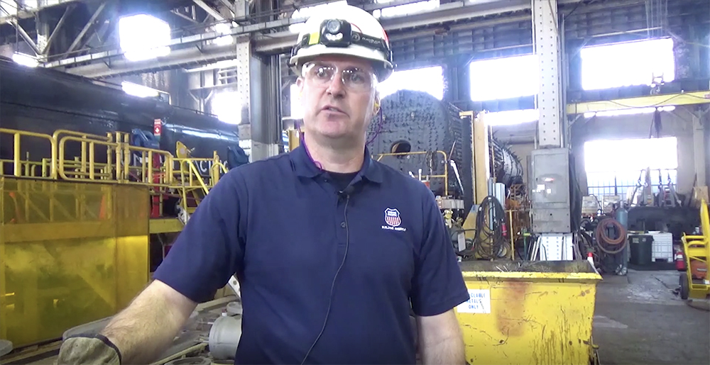 Man with hardhat looking to interviewer in large industrial workshop.