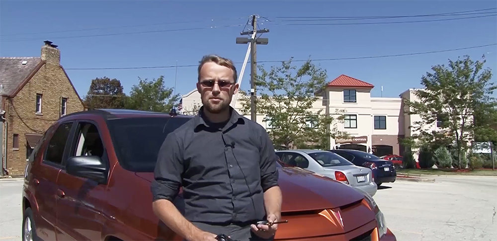 Host Drew Halverson appears in front of a metallic orange Pontiac Aztec on a clear blue-sky day.