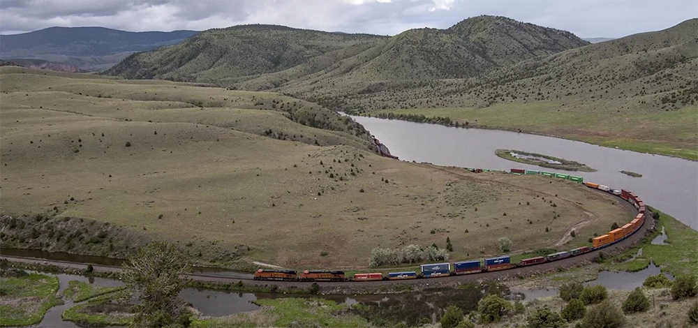 High elevation view of train snaking along banks of large river and distant mountains.