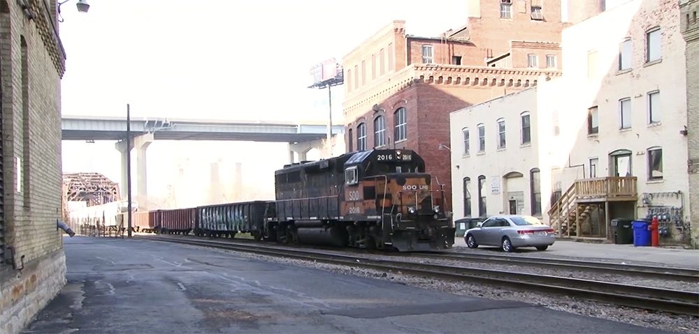 A diesel locomotive in shadow, leads a train through city scenes.