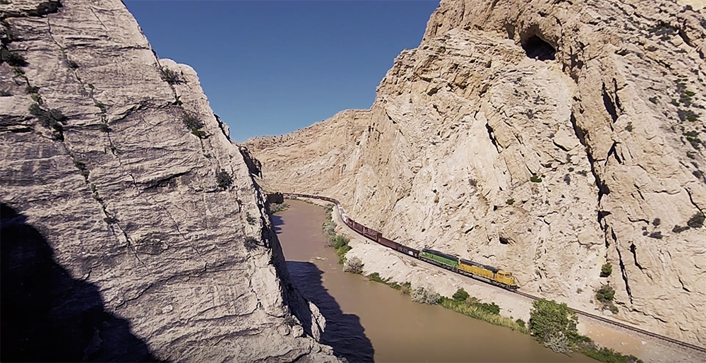 Freight train snaking along the bottom of a rocky canyon just above a river.