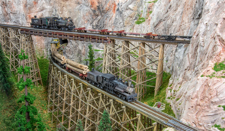 Two model trains powered by steam locomotives traveling across tall bridges