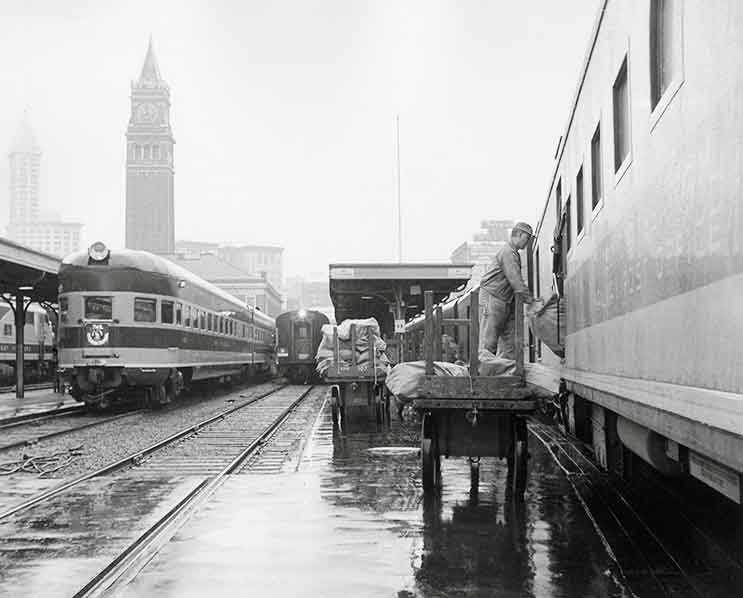 Worker loads bags into a passenger car amid several passenger trains at a station with a tall clock tower.