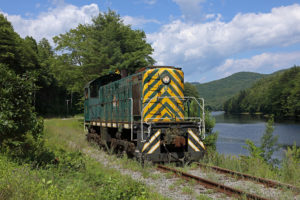 An older green-painted diesel locomotive sits on grassy tracks beside a river in a pastoral summer scene.