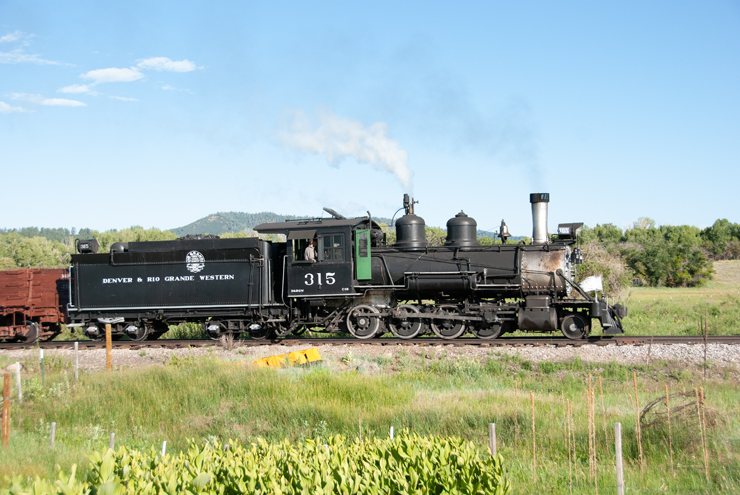 Older steam locomotive pulling a train along a grassy flat area.