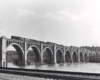 A steam locomotive leads a freight train over a river on a large stone or concrete arch bridge.
