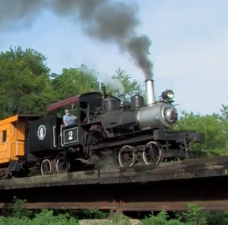 Geared steam locomotive leading a train across a bridge.