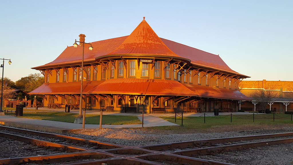 Rounded, old-style passenger train station in low-angle sunlight at a track diamond.