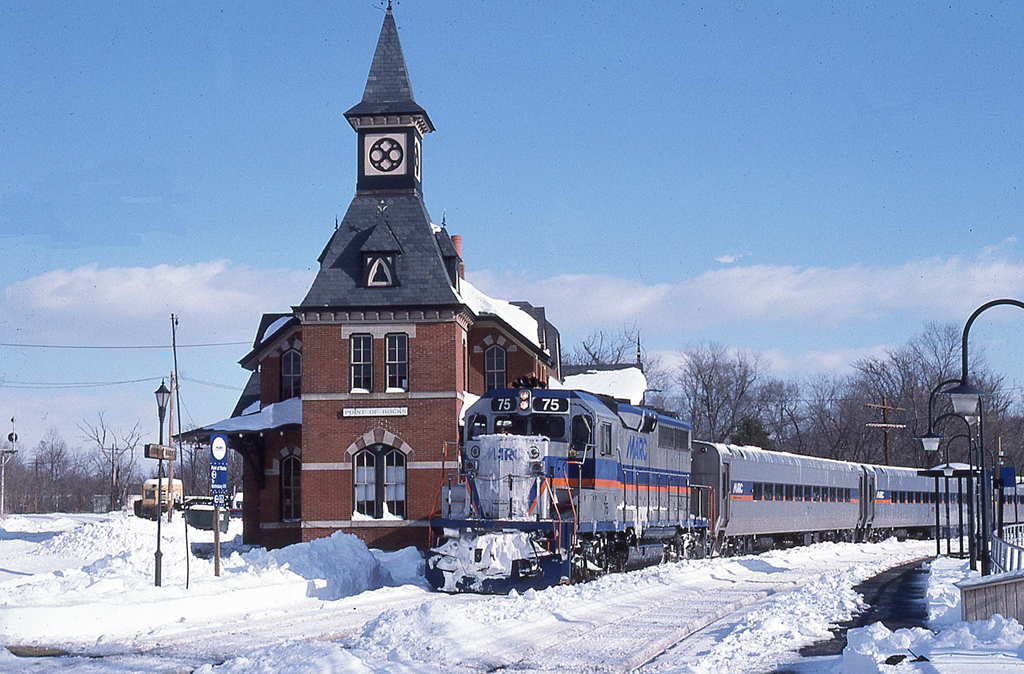 Silver-colored commuter train passes an old-looking passenger train station in the snow.
