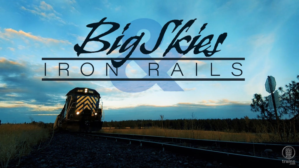 Train moving along the tracks at dusk and Big Skies & Iron Rails logo.
