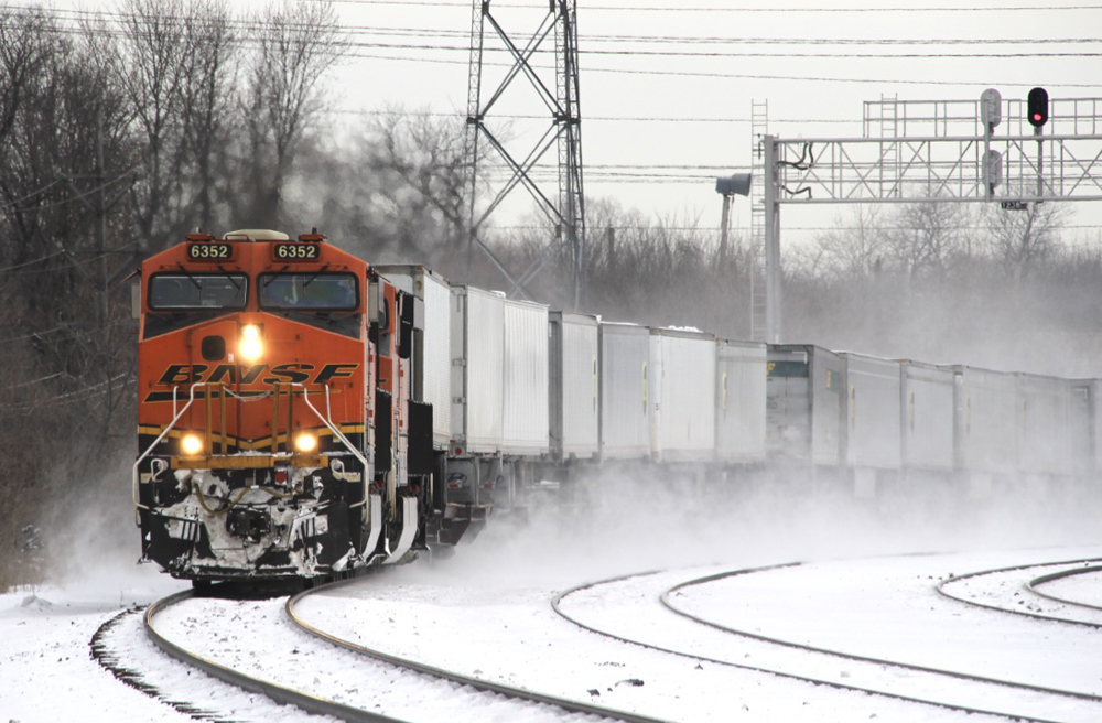 An orange BNSF Railway locomotive leads a train over a snow-covered multi-track main line.