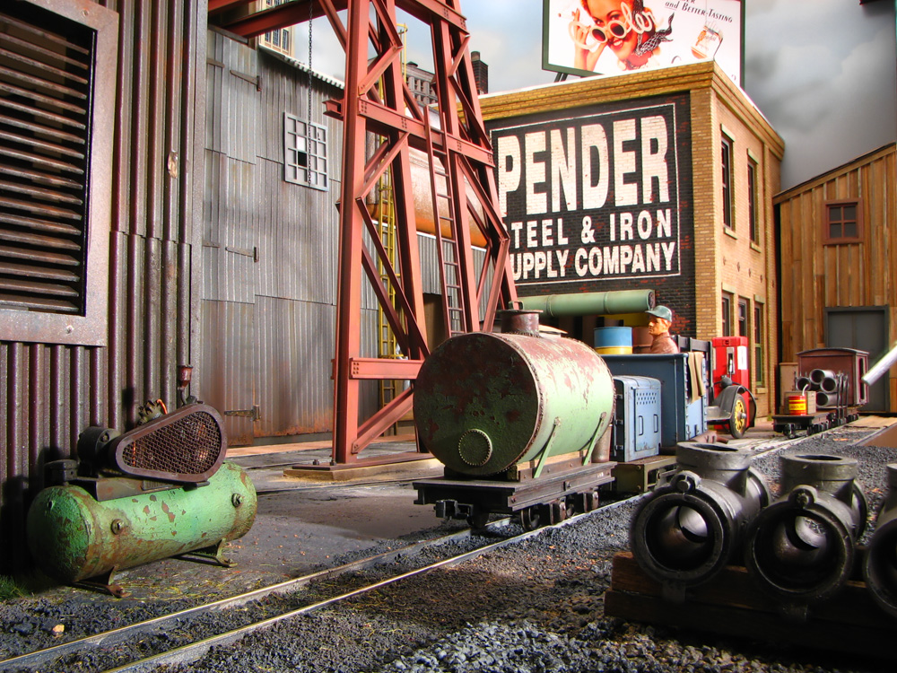 A man on a small industrial shifter moving a tank car on narrow gauge tracks is dwarfed by surrounding industrial buildings