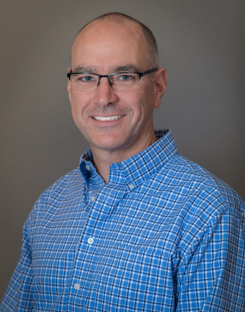 Man with shaved head wearing eyeglasses and blue open-collared shirt.