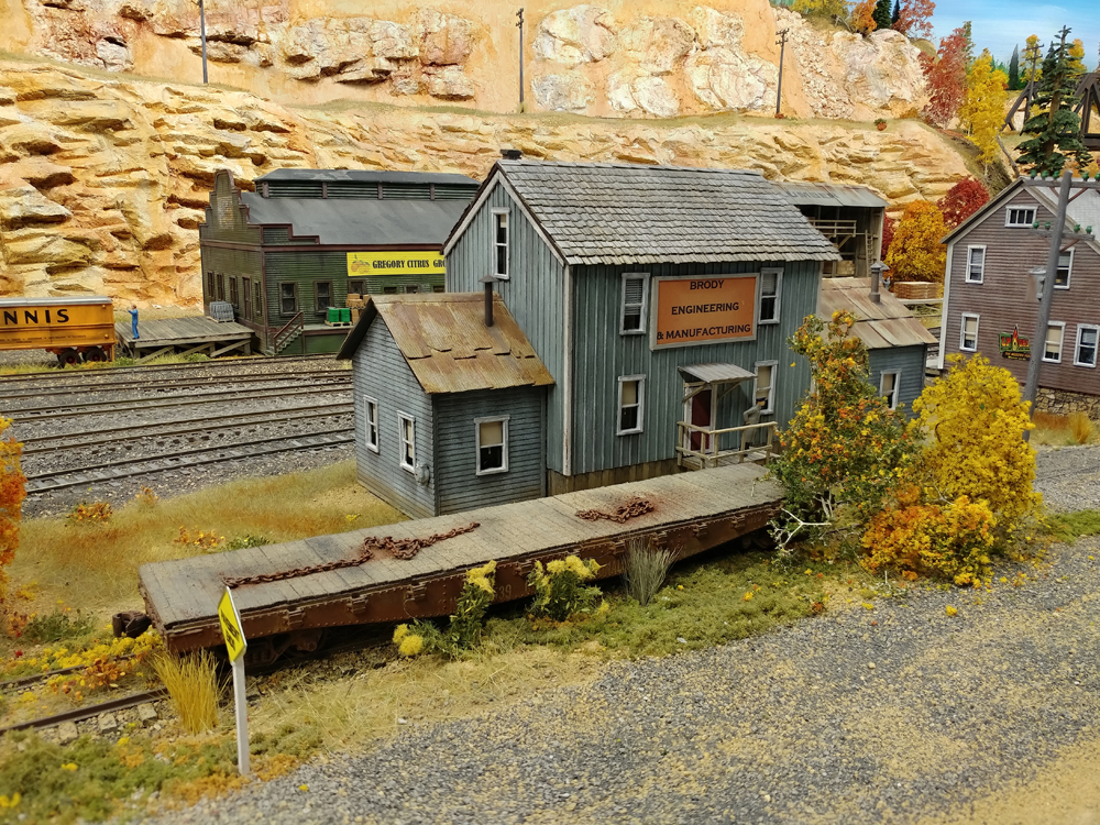 Gray board-and-batten two-story building with abandoned flatcar in foreground, main line running behind