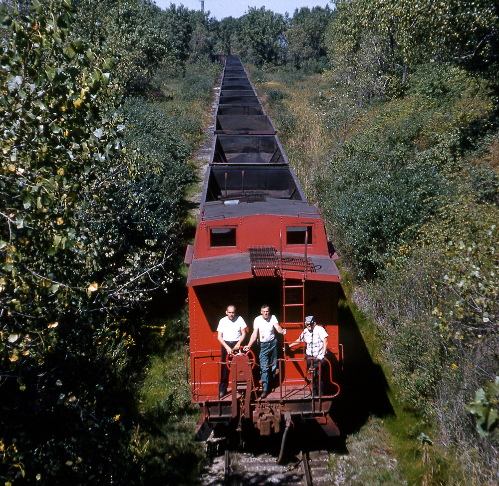 Railroaders on the rear platform of a red caboose behind a long train. Color photograph.