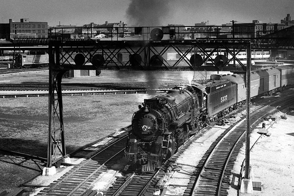 Steam locomotive with passenger train paused underneath a signal bridge in a black and white image.