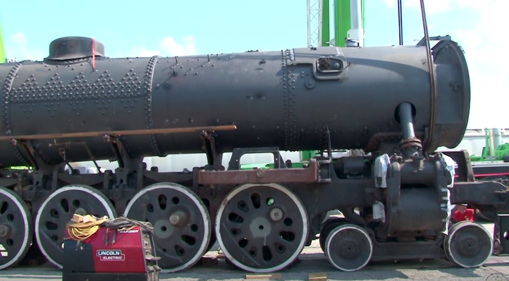 A crane hoists a rusty steam locomotive boiler and frame off of wheel sets on a clear day.