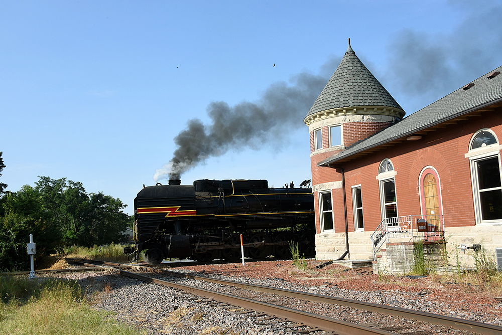 Steam locomotive noses past an old brick train station.