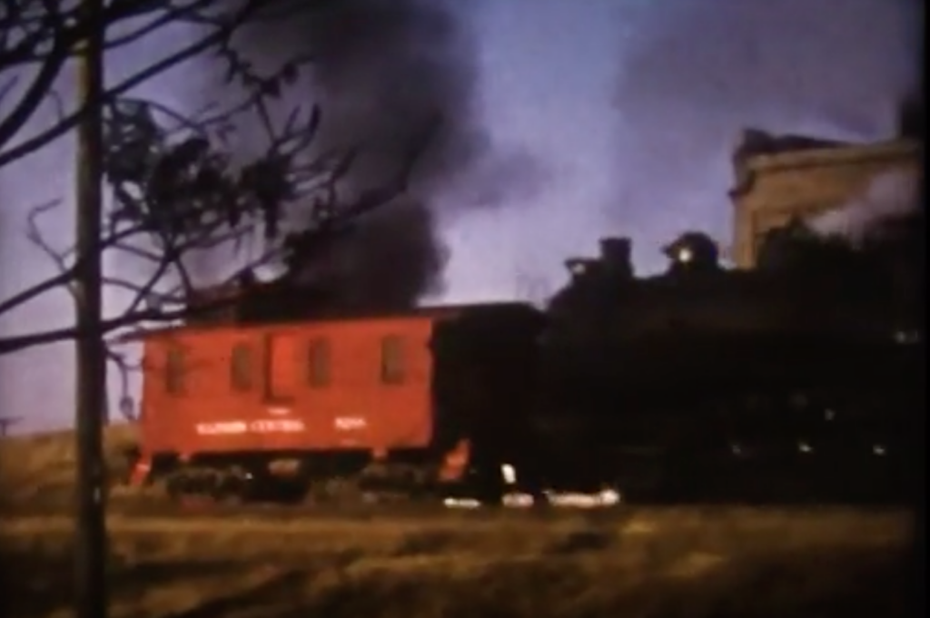 Black steam locomotive pushes a red caboose in low angle sunlight.