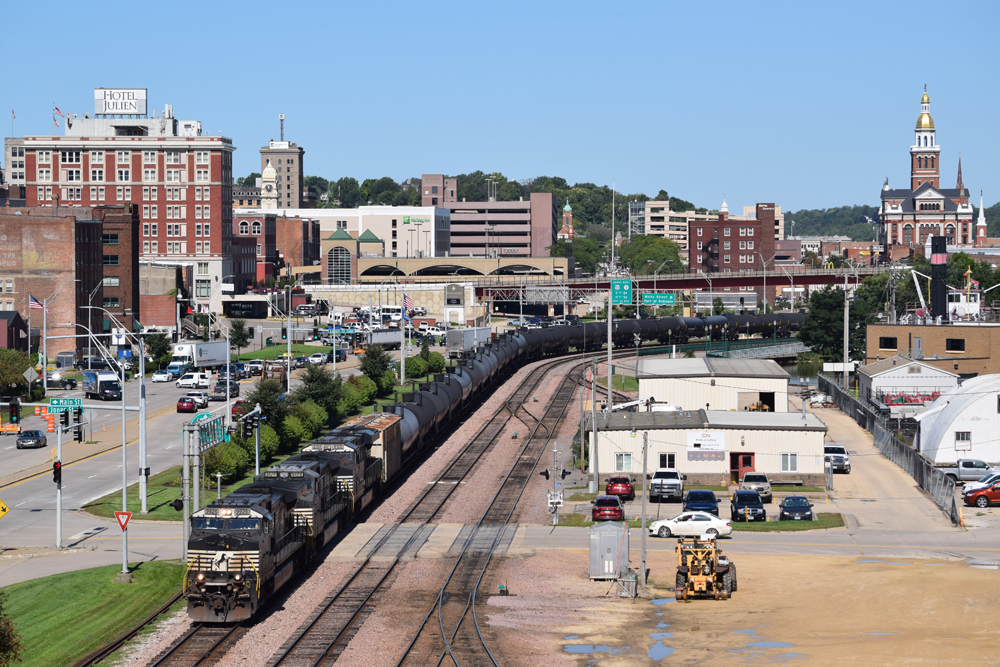 Unit train of tank cars rounds curve in area surrounded by buildings