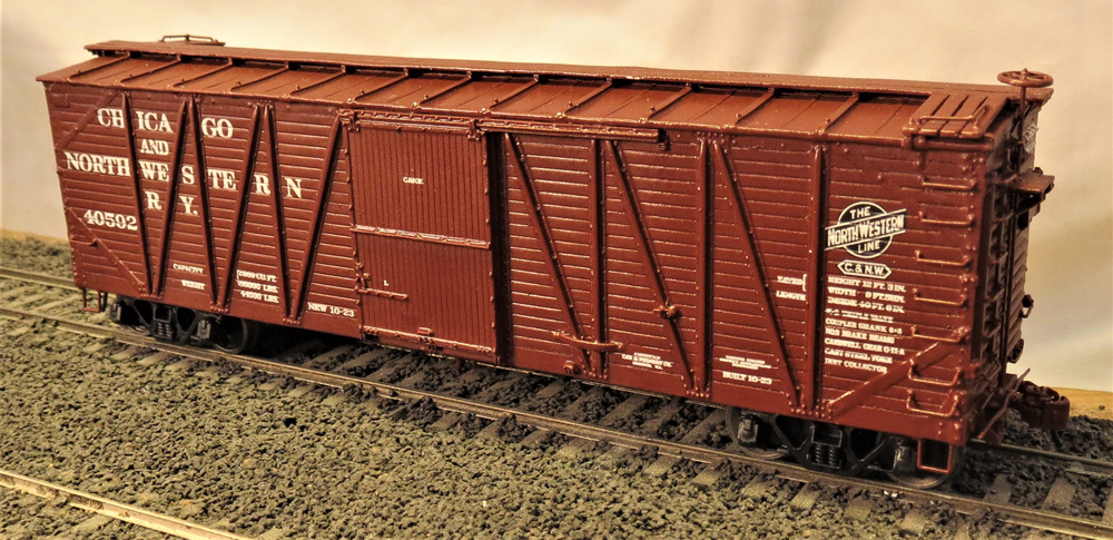 Photo of outside-braced boxcar painted brown on ballasted stretch of track.