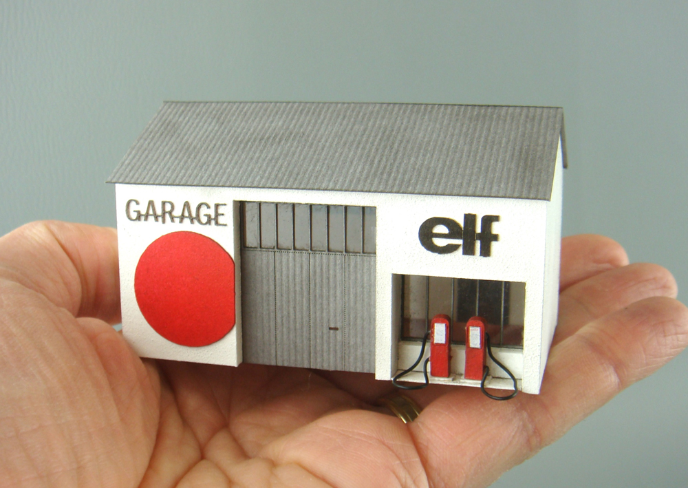 Photo showing N scale Elf gas station painted white, gray, and red with large red circle and two gas pumps in man’s hand.