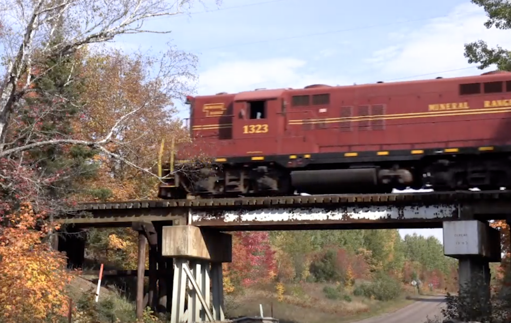 Mineral Range railroad of Michigan:Rust red colored locomotive crosses a bridge over a road in an autumnal scene.