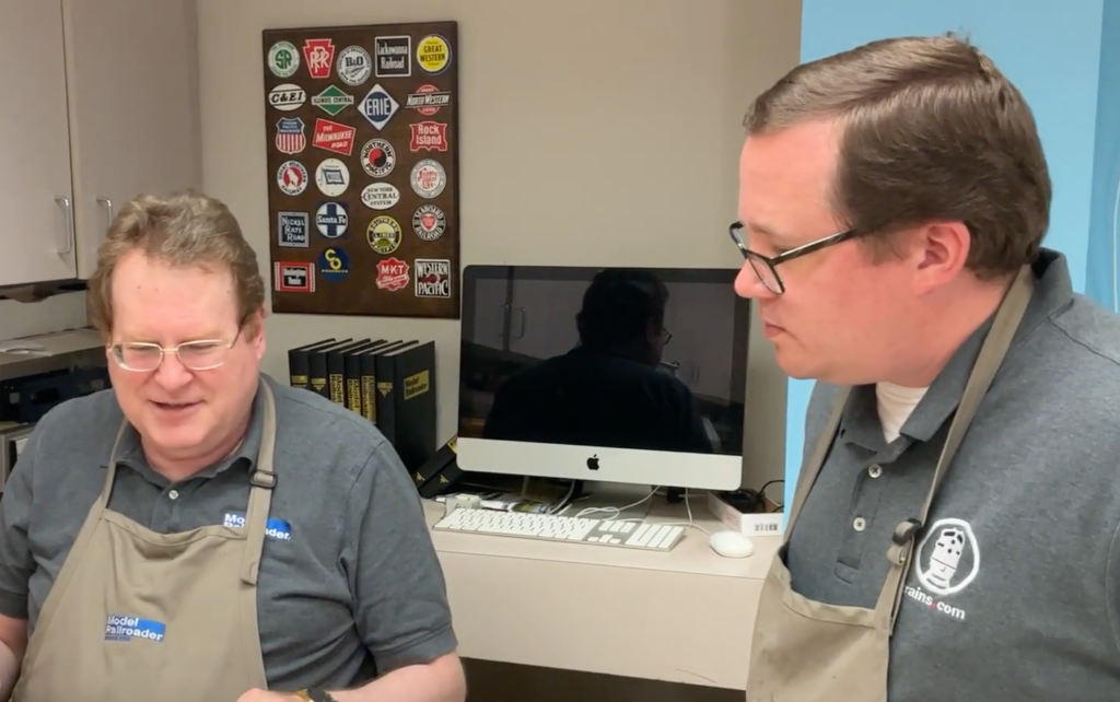 Two men wearing aprons talk.