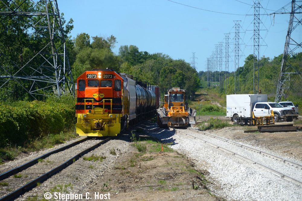 Train led by orange locomotive passes maintenance equipment working on new siding