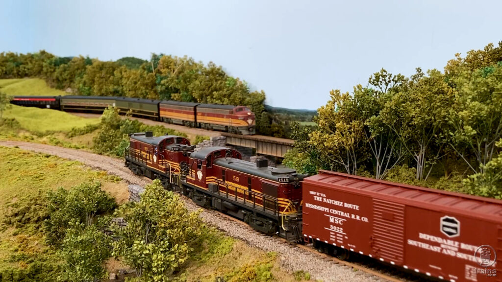Screen shot from a video of two maroon-and-yellow diesels pulling trains through a verdant HO scale landscape on John Callahan’s HO scale East Berkshire Branch