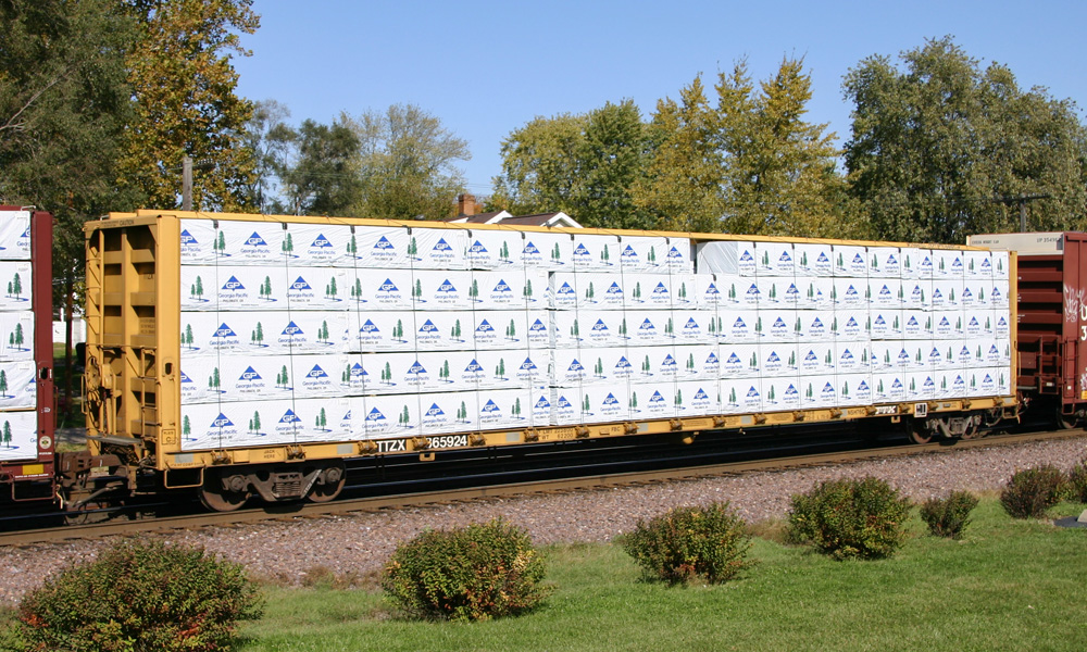 A centerbeam flatcar loaded with white-wrapped bundles of lumber sits in a train