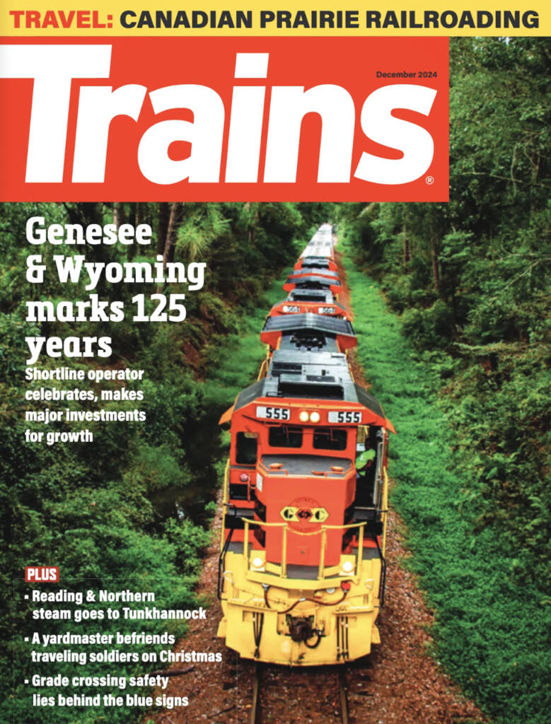 The December 2024 cover of Trains magazine, which features a red and yellow locomotive moving towards the photographer in a forested setting with lush greenery on both sides of the train