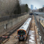 Rapid transit train operates on open-air track with city skyline in distance