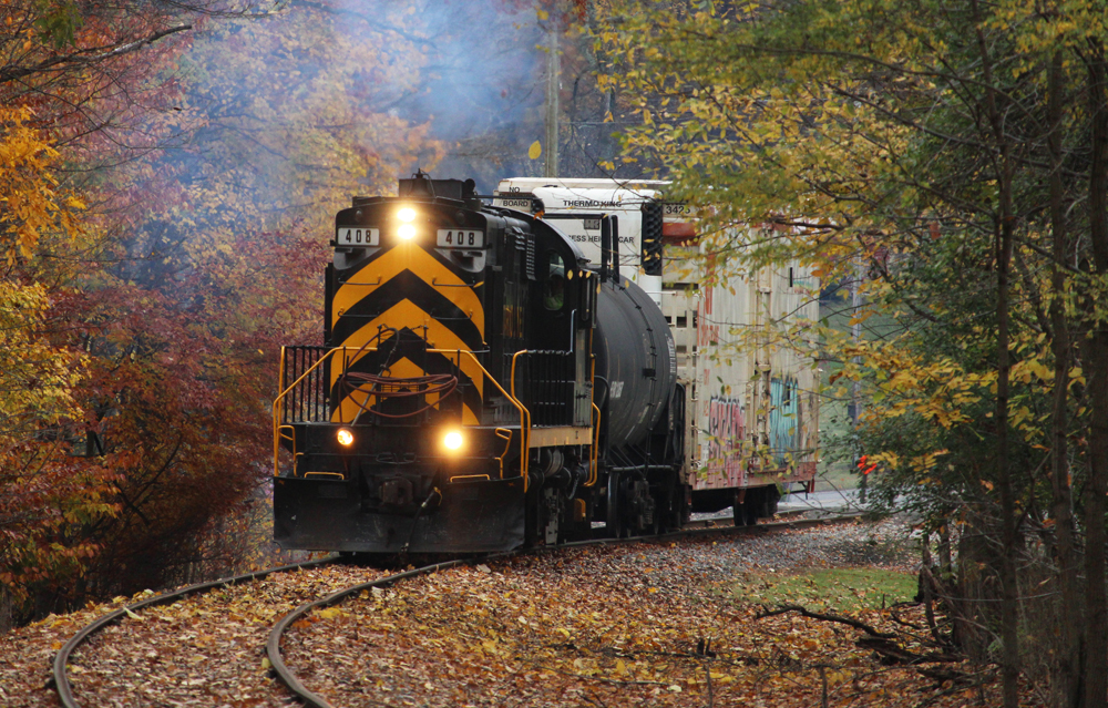Black and orange locomotive with one freight car amid fall foliage