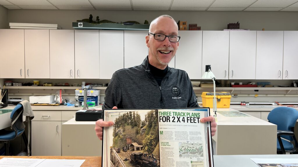 Man shows off book on a model railroad workbench.