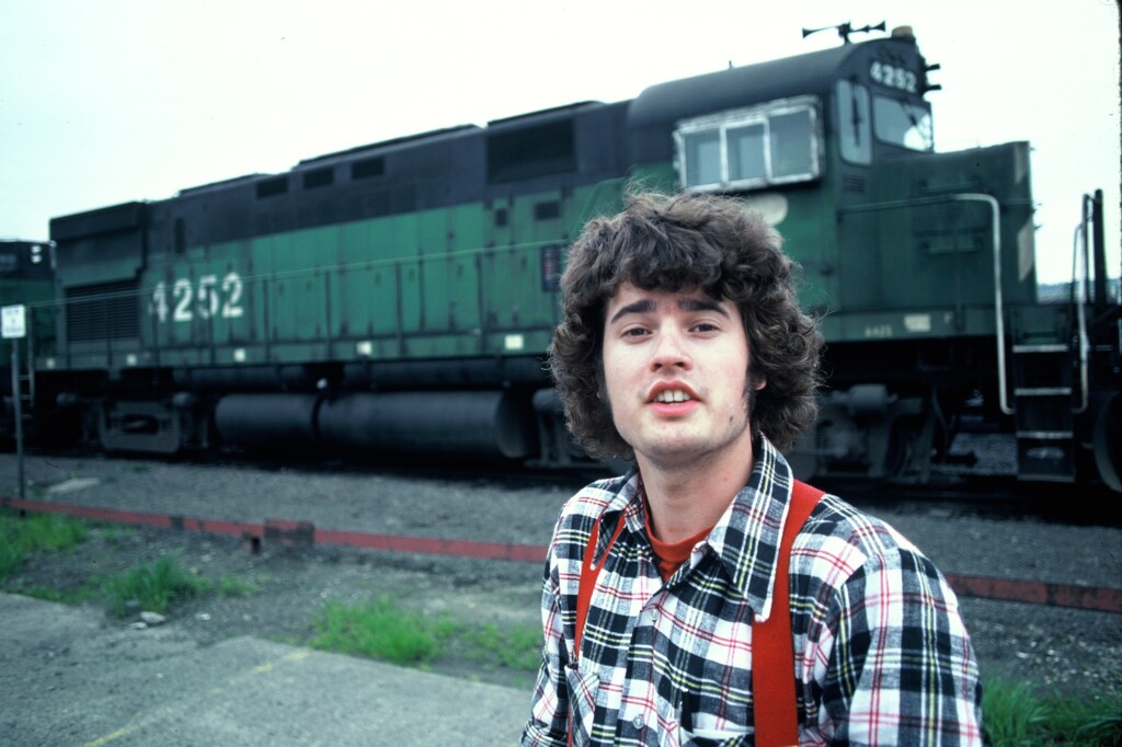 An image showing a young man in a plade shirt and red suspenders standing in front of a green diesel locomotive