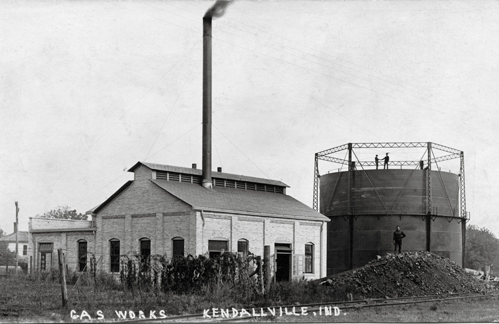 A vintage black-and-white photo of a small industrial brick building with a smokestack, a coal pile outside, and a gas tank in the background