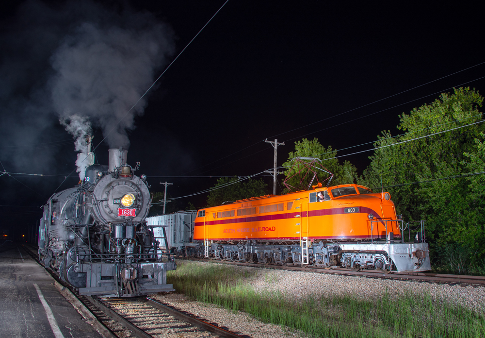 Steam locomotive and orange double-cab electric locomotive posed at night